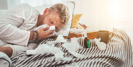 Unhealthy and tired man sneezing or wiping snotty nose man lying in a cozy home bed beside a lot of used paper tissues and medicines on the blanket. Season virus flu and home quarantine concept image.の写真素材