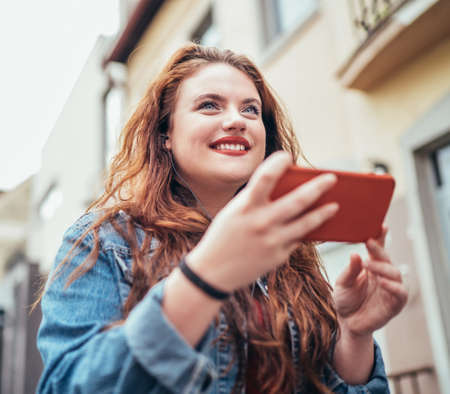 Red curled long hair caucasian teen girl portrait walking on the street and browsing the internet using the modern smartphone. Modern people with technology devices concept image.の写真素材