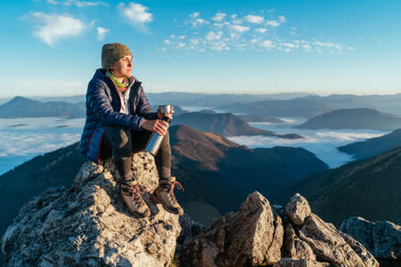 Young hiker woman sitting on the mountain summit cliff, drinking tea from a flask and enjoying mountains valley covered with clouds view. Successful summit climbing concept image.の写真素材