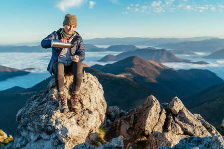 Active backpacker hiker woman sitting on the mountain summit cliff, pouring a tea from a flask and enjoying mountains valley covered with clouds view. Successful summit climbing concept image.の写真素材