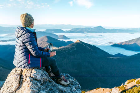 Young hiker woman sitting on the mountain summit cliff and enjoying mountains valley covered with clouds view. Successful summit climbing concept image.の写真素材