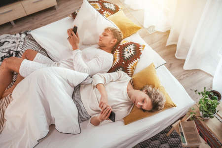Top view of the couple lying in a cozy bed in bedroom and browsing internet and checking mails using each other modern smartphone. Modern technology, social issues and relations problems concept imageの写真素材