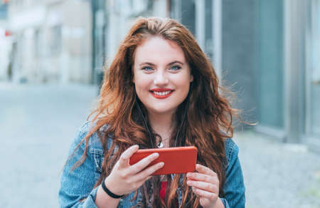Portrait of smiling red curled long hair caucasian teen girl walking on the street and video chatting using the modern smartphone. Modern people with technology devices concept image.の写真素材