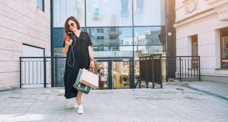 Smiling beautiful modern middle-aged female Portrait dressed black dress and sunglasses with shopping bags browsing slim smartphone listening music with wired earphones. Beautiful people concept imageの写真素材