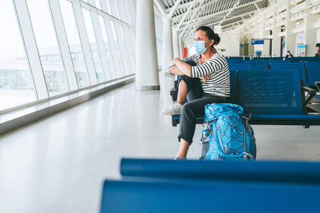 Lonely female solo traveler with backpack sitting in the empty airport passenger transfer hall in protective face mask and looking out large windows. Traveling in worldwide pandemic time concept imageの写真素材