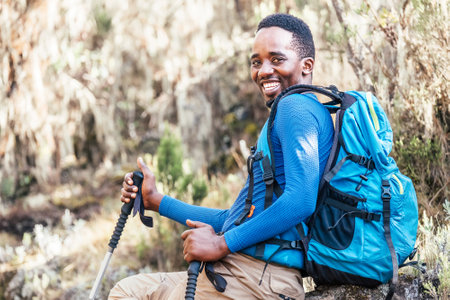 Portrait of a cheerfully smiling African-American Ethnicity young man sitting with a backpack and trekking poles and resting in the forest during a hiking walk. Active people and sport concept.の写真素材
