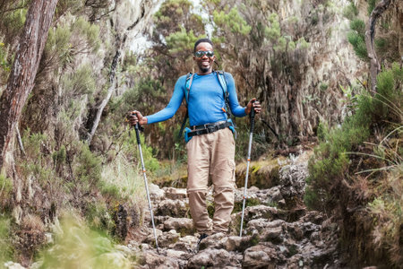 Portrait of a cheerfully smiling African-American Ethnicity young man in sunglasses. He having a walk with a backpack using trekking poles to Mweka gate, Kilimanjaro. Active climbing people concept.の写真素材