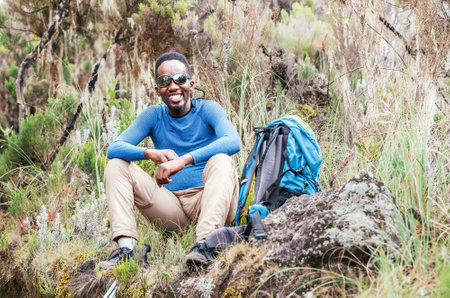 Portrait of a cheerfully smiling African-American Ethnicity young man in sunglasses sitting with a backpack, resting in the forest and looking at the camera. Active people and traveling concept.の写真素材