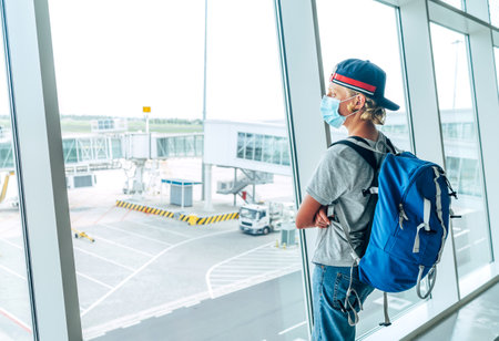 Portrait of lonely teen solo traveler with a backpack in empty airport passenger transfer hall in protective face mask and looking out large windows. Traveling in worldwide pandemic time concept imageの写真素材
