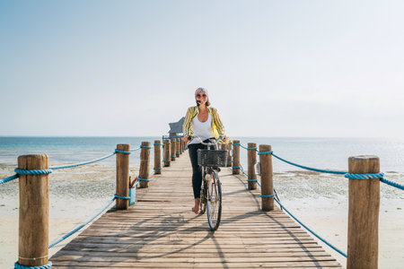 Portrait of a happy smiling woman dressed in light summer clothes and sunglasses riding a bicycle on the wooden sea pier and looking at camera. Careless vacation in tropical countries concept imageの写真素材