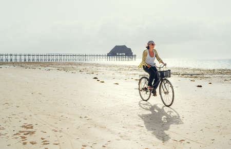 Middle-aged female dressed light summer clothes riding old vintage bicycle with front basket on the lonely low tide ocean white sand coast on Kiwengwa beach on Zanzibar island, Tanzania.の写真素材