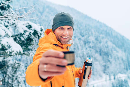 A middle-aged man offering a hot drink cup from a flask dressed in a bright orange softshell jacket while he trekking the winter mountains route. Active people in the nature concept image.の写真素材