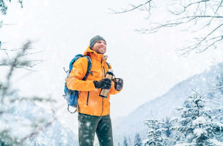 Smiling Man under snowfall dressed bright orange softshell jacket with a hot drink flask while he...