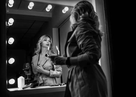 Sensual portrait of middle-aged seductive woman with red wine glass gazing at mirror reflection appreciating her Trench coat outfit. Human emotion or clothing fashion concept black and white image.の写真素材
