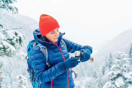 Smiling Woman dressed warm down jacket pouring a hot drink from flask to mug while she has a break on trekking winter mountains route. Active people in the nature concept image.の写真素材