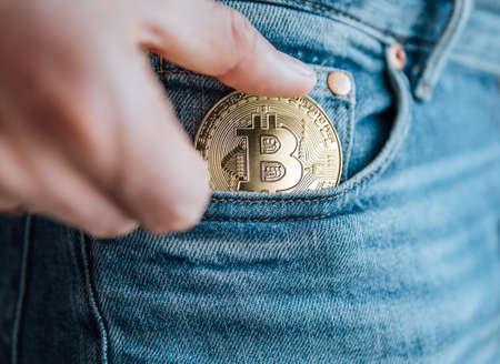 Close up footage of man's fingers putting the Bitcoin coin in the blue jeans small pocket. Savings, digital currency, cryptocurrency, investing, and smart outgoings managing concept.の写真素材