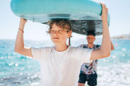 Blonde teenage son with father surfers smiling carrying the stand-up paddleboard with a paddle on the bright sunny day noon. Active family summer vacation time near the sea concept image.の写真素材