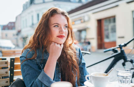 Portrait of sad red curled long hair caucasian woman sitting on a cozy outdoor cafe terrace on the street and looking at the street. Young woman taking a break in her city bicycle tour.の写真素材