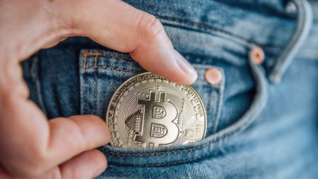 Close up footage of man's fingers putting the Bitcoin coin in the blue jeans small pocket. Savings, digital currency, cryptocurrency, investing, and smart outgoings managing concept.の写真素材