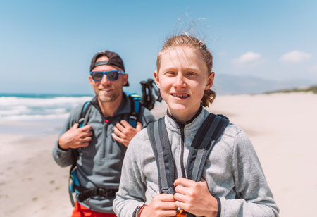 Father with teenager son with backpacks walking by the sandy seaside beach. They smiling and looking at the camera. Active happy family people vacation time concept image.の写真素材