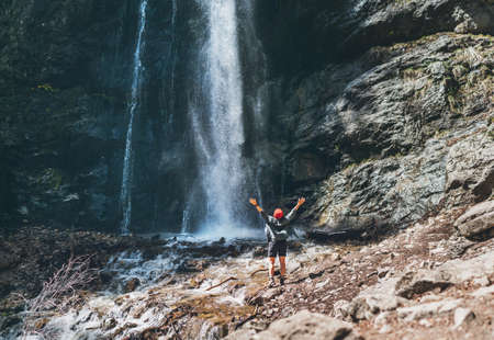 A woman dressed in a red hat and active trekking clothes standing near the mountain river waterfall rose arms up and enjoying the splashing Nature power. Traveling, trekking, and nature concept imageの写真素材