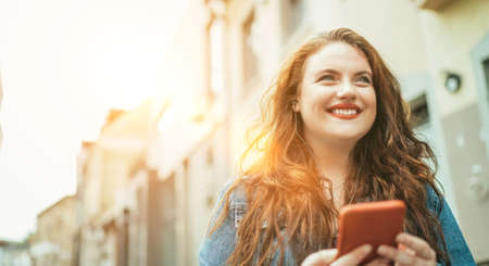 Smiling red curly long hair caucasian teen girl walking on the street and browsing the internet using the modern smartphone. Modern people with technology devices concept image.の写真素材