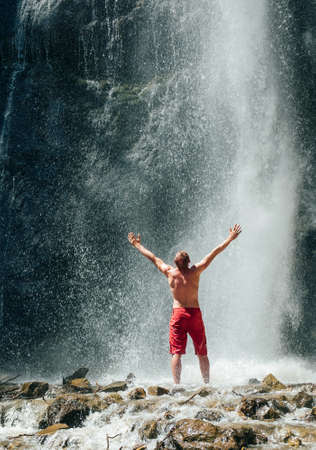 Middle-aged man dressed only red trekking shorts standing under the mountain river waterfall, rose arms up and enjoying the splashing Nature power. Traveling, trekking and nature concept image.の写真素材