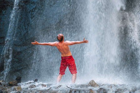 Shirtless Man dressed only red trekking shorts standing under the mountain river waterfall, spread his arms and enjoying the splashing Nature power. Traveling, trekking and nature concept image.の写真素材