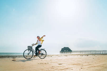 Kiwengwa beach on Zanzibar island, Tanzania.Young female dressed light summer clothes joyfully threw up her hand riding old vintage bicycle with front basket on the low tide ocean white sand coast onの写真素材
