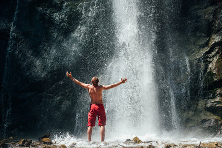 Middle-aged man dressed only red trekking shorts standing under the mountain river waterfall, rose arms up and enjoying the splashing Nature power. Traveling, trekking and nature concept image.の写真素材