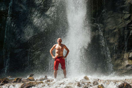 A smiling middle-aged topless man standing under the mountain river waterfall and enjoying the splashing Nature power. Fit people, trekking, and a natural beauty concept image.の写真素材