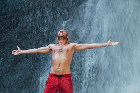 Middle-aged man dressed only in red trekking shorts standing under the mountain river waterfall spread arms and enjoying the splashing Nature power. Traveling, trekking and nature concept image.の写真素材