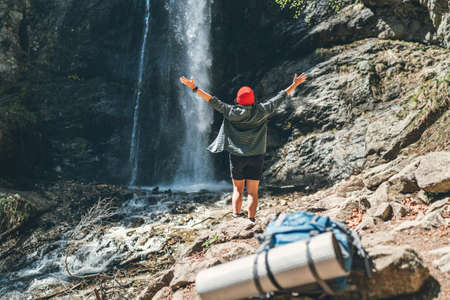 Woman dressed in red hat and active trekking clothes near the mountain river waterfall rose arms up and enjoying the Nature. BAckpack on the foreground.Traveling, trekking, and nature concept imageの写真素材