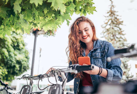 Portrait of smiling red curled long hair caucasian teen girl block out bike at Bicycle sharing point using the modern smartphone. Green urban transport and modern technology devices concept image.の写真素材
