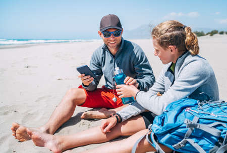 Teenager Boy passing drinking water bottle to Smiling Father in sunglasses with smartphone sitting with backpacks on the sandy seaside beach. Active happy family people vacation concept imageの写真素材