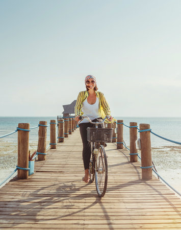 Portrait of a happy smiling woman dressed in light summer clothes and sunglasses riding a bicycle on the wooden sea pier and looking at camera. Careless vacation in tropical countries concept imageの写真素材
