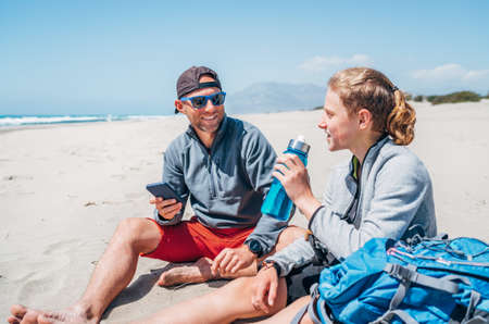 Smiling Father in sunglasses with smartphone sitting with teenager son with backpacks on the sandy seaside beach. Boy drinking water from bottle. Active happy family people vacation time concept imageの写真素材