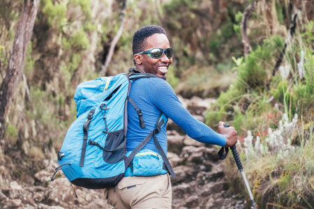 Portrait of a cheerfully smiling African-American Ethnicity young man in sunglasses. He having a walk with a backpack using trekking poles to Mweka gate, Kilimanjaro. Active climbing people conceptの写真素材