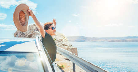 Cheerful Woman portrait enjoying the seaside road trip. Dressed a black dress with straw hat and sunglasses she wide opened arms and shining with happiness. Summer vacation traveling by auto concept.の写真素材