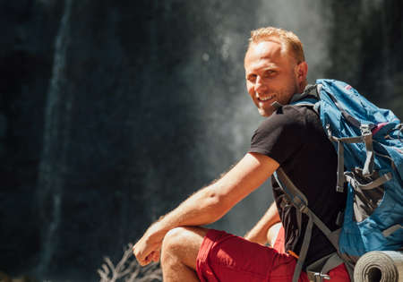 Sincerely smiling Man portrait with backpack dressed in active trekking clothes sitting near mountain river waterfall smiling and enjoying the Nature. Traveling, trekking, nature concept imageの写真素材