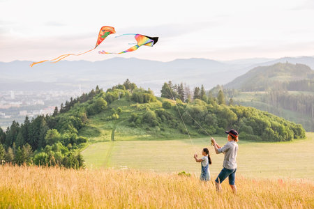 Kids playing: sister with a brother with launching colorful kites - popular outdoor toy on the high grass hills meadow. Happy childhood moments or outdoor time spending concept image.の写真素材