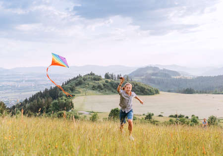 Smiling teenager boy with flying colorful kite on the high grass meadow in the mountain fields. Happy childhood moments or outdoor time spending concept image.の写真素材