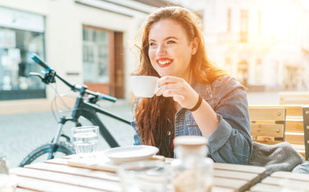 Portrait of sad red curled long hair caucasian teen girl sitting on a cozy cafe outdoor terrace on the street and drinking coffee. Young woman taking a break in her city bicycle tour.の写真素材