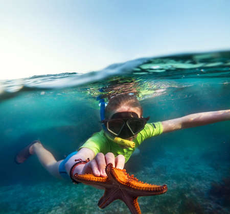 Underwater shot of a young teenage boy snorkeling with dive face mask in the blue Indian Ocean waves near the Zanzibar island with a big orange starfish in his hand. Exotic vacations concept image.の写真素材