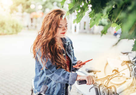 Portrait of smiling at camera red curled hair caucasian teen girl unlocking bike at Bicycle sharing point using the modern smartphone. Green urban transport and modern technology devices concept imageの写真素材
