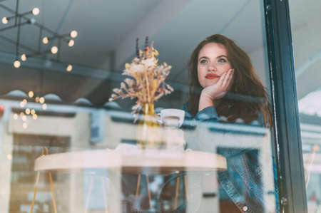 Portrait of sad red curled long hair caucasian girl sitting at table in cozy cafe and looking through the window glass at the street.の写真素材