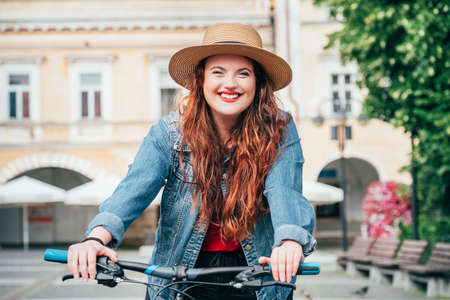 Portrait of smiling long red curly hair caucasian teen girl in a straw hat riding a vintage bicycle in the old town center. Eco transport sharing or city bike hiring concept image.の写真素材