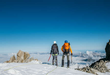 Couple connected team rope  with climbing harness dressed mountaineering clothes with backpacks and ice axes enjoying views ascending Mont Blanc (Monte Bianco) summit near Aiguille du Midi, France.の写真素材