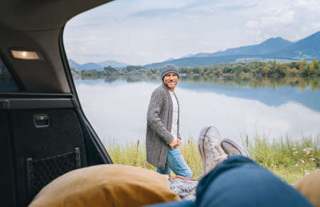 Smiling middle-aged man dressed in warm knitted clothes and jeans near the car. Trunk mountain lake view with woman legs in white sneakers. Cozy early autumn couple auto traveling concept image.の写真素材