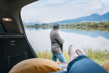 Car trunk view with woman legs in white sneakers on the man dressed warm knitted clothes and jeans enjoying the mountain lake view. Cozy early autumn couple auto traveling concept image.の写真素材